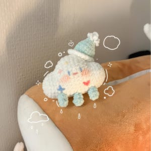 Puede incluir: Un juguete de peluche de nube blanca de crochet con un sombrero azul y una cara sonriente. El juguete está sentado sobre una superficie marrón.