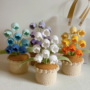 Op de afbeelding: Gehaakte bloemstukken in beige potten. Elke pot bevat bloemen in verschillende kleuren: blauw, paars, wit en geeloranje. De bloemen hebben groene bladeren en zijn decoratief gerangschikt. De potten hebben een geschulpte rand.