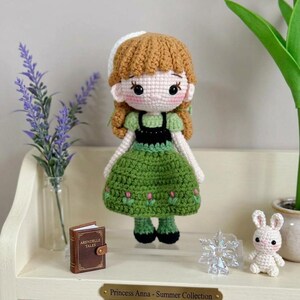 Peut inclure: Poup&eacute;e crochet&eacute;e d'une princesse v&ecirc;tue d'une robe verte avec des d&eacute;tails de fleurs roses, un corsage noir et des cheveux bruns. La poup&eacute;e a un bonnet blanc et fait partie de la "Collection &Eacute;t&eacute;". Un petit livre, un flocon de neige et un lapin crochet&eacute; sont sur l'&eacute;tag&egrave;re.