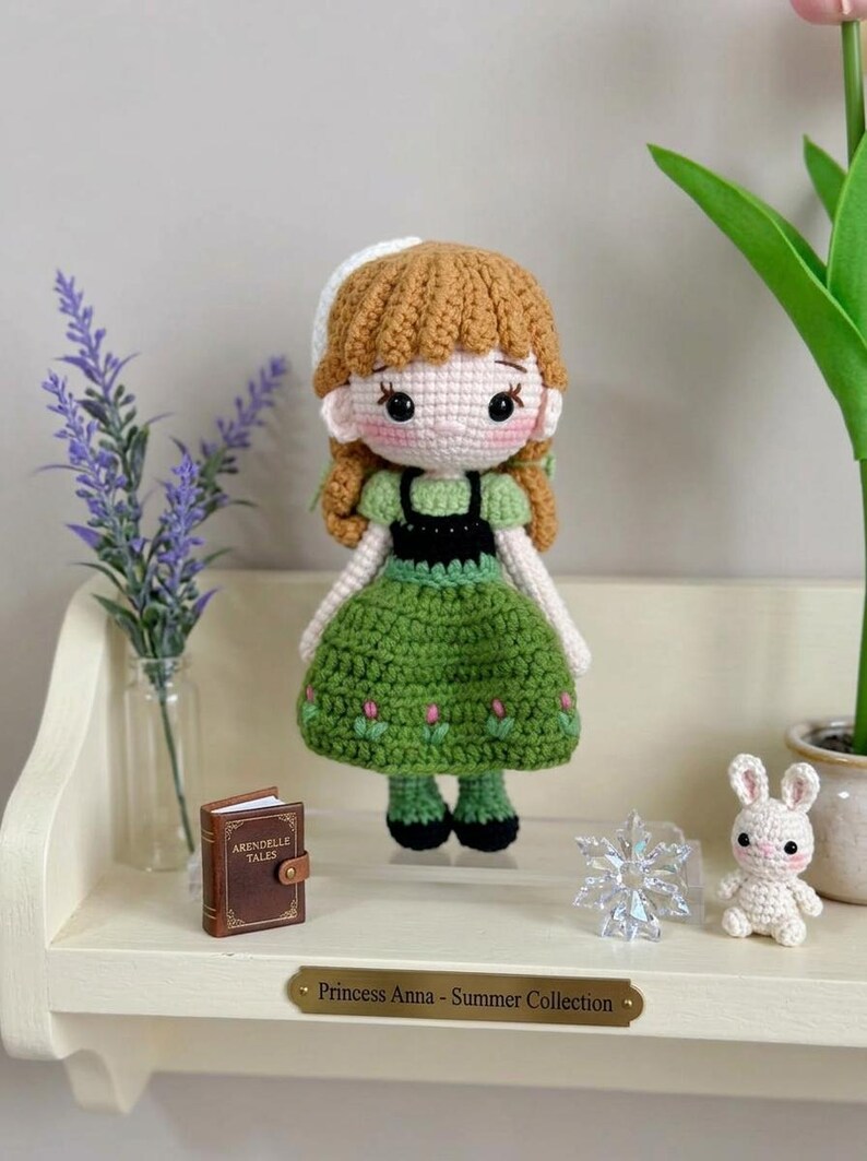 Peut inclure: Poup&eacute;e crochet&eacute;e d'une princesse v&ecirc;tue d'une robe verte avec des d&eacute;tails de fleurs roses, un corsage noir et des cheveux bruns. La poup&eacute;e a un bonnet blanc et fait partie de la "Collection &Eacute;t&eacute;". Un petit livre, un flocon de neige et un lapin crochet&eacute; sont sur l'&eacute;tag&egrave;re.