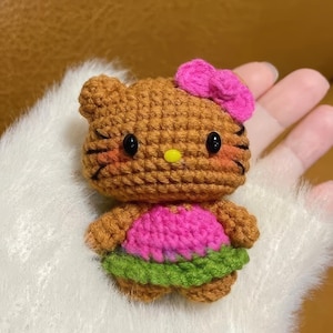 Peut inclure: Poup&eacute;e au crochet faite &agrave; la main en forme de chat de dessin anim&eacute;. La poup&eacute;e a un corps marron, un n&oelig;ud rose et une robe rose et verte. La poup&eacute;e a des yeux noirs et un nez jaune.