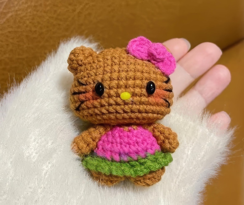 Peut inclure: Poup&eacute;e au crochet faite &agrave; la main en forme de chat de dessin anim&eacute;. La poup&eacute;e a un corps marron, un n&oelig;ud rose et une robe rose et verte. La poup&eacute;e a des yeux noirs et un nez jaune.