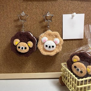 Könnte beinhalten: Zwei Schlüsselanhänger mit Teddybären aus Plüsch mit sternförmigen Verschlüssen. Einer ist braun mit gelben Akzenten, der andere ist beige mit rosa Akzenten. Ein kleiner bärenförmiger Artikel befindet sich in einem gelben Korb.