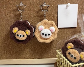 May include: Two plush bear keychains with star-shaped clasps. One is brown with yellow accents, the other is tan with pink accents. A small bear-shaped item sits in a yellow basket.