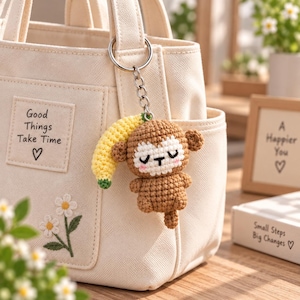 May include: A cream-coloured canvas tote bag featuring an embroidered daisy and a square patch with the text Good Things Take Time and a heart. A crocheted monkey keychain with a yellow banana is attached to the silver metal ring on the bag.