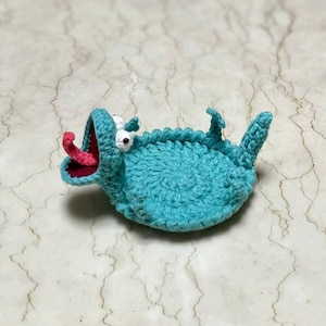 Puede incluir: Criatura de ganchillo turquesa hecha a mano, con la boca muy abierta y una lengua rosa. La criatura tiene ojos blancos y aletas pequeñas. El trabajo de ganchillo es detallado y el diseño es caprichoso y juguetón.