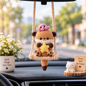 May include: A crocheted otter hanging ornament suspended from a beige rope swing. The brown and cream otter holds a yellow star, with a small pink octopus on its head. The ornament features dark brown paws and a long tail, crafted with yarn.