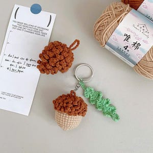 May include: Crocheted acorn and pinecone keychains in shades of brown and beige, with a green leaf accent. A ball of beige yarn and a handwritten note are also in the image. The keychains are handmade.