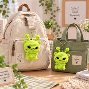 May include: A beige canvas backpack and a sage green canvas tote bag, each featuring a bright lime green crocheted alien keychain. The backpack has a patch reading Good Things Take Time, and the tote has a patch reading Live A Kinder Day.