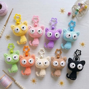 May include: A collection of nine crocheted cat keychains in yellow, pink, purple, light blue, green, orange, cream, tan, and black. Each cat features large white eyes with black pupils, a small embroidered mouth, and a star-shaped plastic clasp.