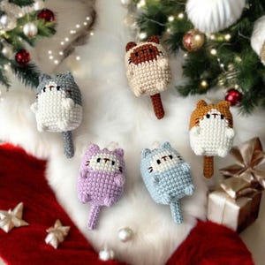 May include: Five crocheted cat ornaments in different colors, including gray, brown, purple, and blue. The ornaments are hanging from a white fuzzy surface.