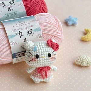 Peut inclure: Une poup&eacute;e Hello Kitty crochet&eacute;e avec un n&oelig;ud et une robe roses, sur fond de pelotes de laine roses et de petites formes crochet&eacute;es. L'emballage du fil comporte des caract&egrave;res chinois. La poup&eacute;e a des yeux noirs et des moustaches.