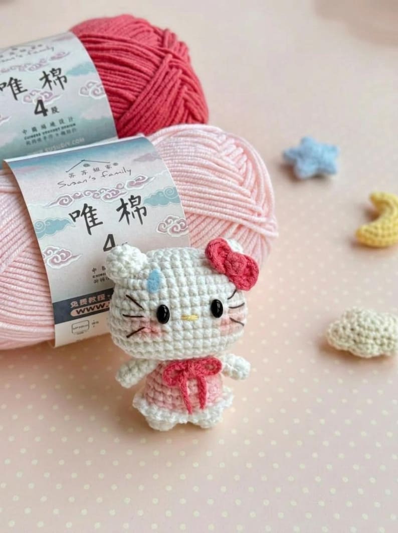 Peut inclure: Une poup&eacute;e Hello Kitty crochet&eacute;e avec un n&oelig;ud et une robe roses, sur fond de pelotes de laine roses et de petites formes crochet&eacute;es. L'emballage du fil comporte des caract&egrave;res chinois. La poup&eacute;e a des yeux noirs et des moustaches.