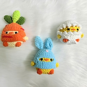 May include: Three handmade crochet plush toys: an orange carrot with green leaves, a blue bunny with a yellow face, and a yellow chick hatching from a white egg. The toys are made with soft yarn and have black eyes and red accents.