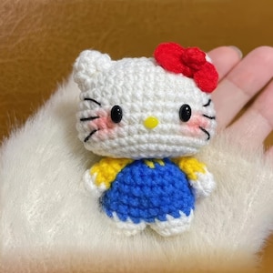 Peut inclure: Une poup&eacute;e au crochet faite &agrave; la main avec une t&ecirc;te blanche, un n&oelig;ud rouge, des yeux noirs et des joues roses. La poup&eacute;e porte une tenue bleue et jaune. La poup&eacute;e est un personnage populaire.