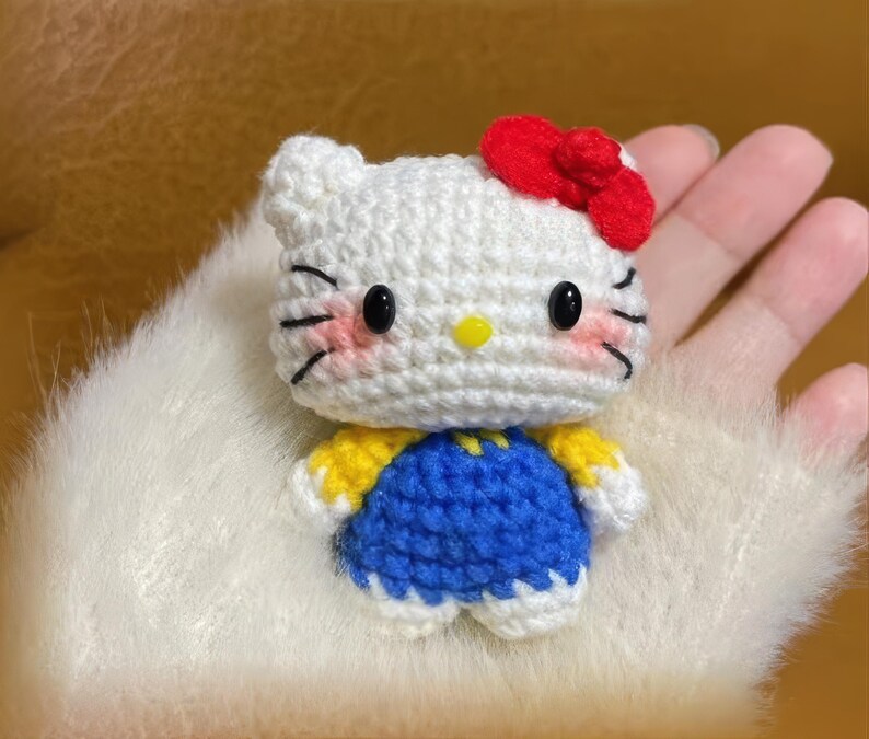 Peut inclure: Une poup&eacute;e au crochet faite &agrave; la main avec une t&ecirc;te blanche, un n&oelig;ud rouge, des yeux noirs et des joues roses. La poup&eacute;e porte une tenue bleue et jaune. La poup&eacute;e est un personnage populaire.