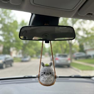 Pu&ograve; includere: Un ornamento a forma di personaggio lavorato all'uncinetto in grigio e bianco, appeso a un cordino marrone. La figura ha due orecchie a punta, un piccolo accento verde sulla sommit&agrave;, occhi a perla neri e una bocca cucita. &Egrave; appeso allo specchietto.