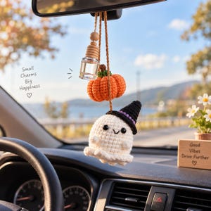 May include: A car hanging ornament featuring a crocheted white ghost wearing a black witch hat with a purple band, a crocheted orange pumpkin, and a small glass essential oil bottle with a wooden cap, all suspended by brown cords.