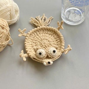 Puede incluir: Un posavasos hecho a mano, de ganchillo beige, con forma de criatura caprichosa con ojos grandes y saltones y pequeños apéndices. El posavasos es redondo y se encuentra sobre una superficie gris claro. Ovillos de lana y un vaso en el fondo.