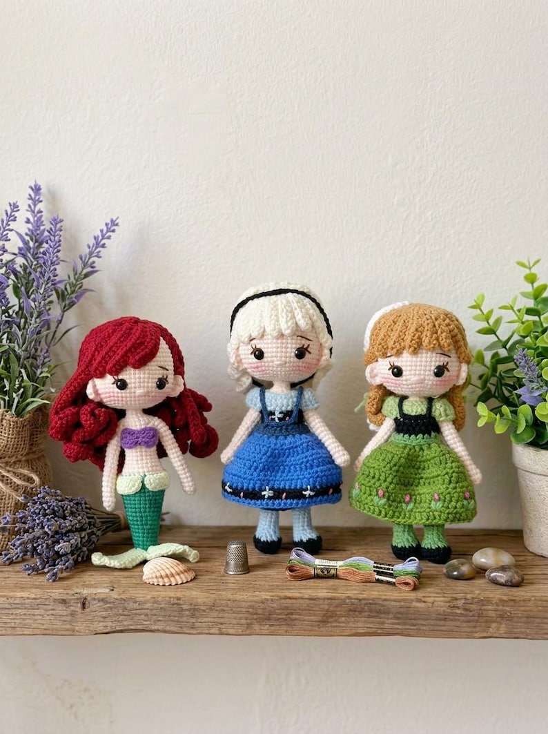 Peut inclure: Trois poup&eacute;es au crochet faites &agrave; la main : une sir&egrave;ne aux cheveux roux, une poup&eacute;e en robe bleue et une poup&eacute;e en robe verte. Les poup&eacute;es sont expos&eacute;es sur une &eacute;tag&egrave;re en bois avec de la lavande, un d&eacute; &agrave; coudre et du fil &agrave; broder.