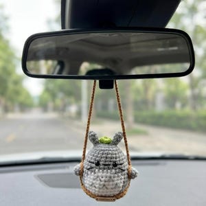 Pu&ograve; includere: Un ornamento all'uncinetto grigio e bianco appeso a un cordino marrone, con piccole orecchie, occhi neri e un germoglio verde sulla testa. La figura &egrave; seduta su un'altalena all'uncinetto marrone con una piccola etichetta rettangolare con scritto HANDMADE.