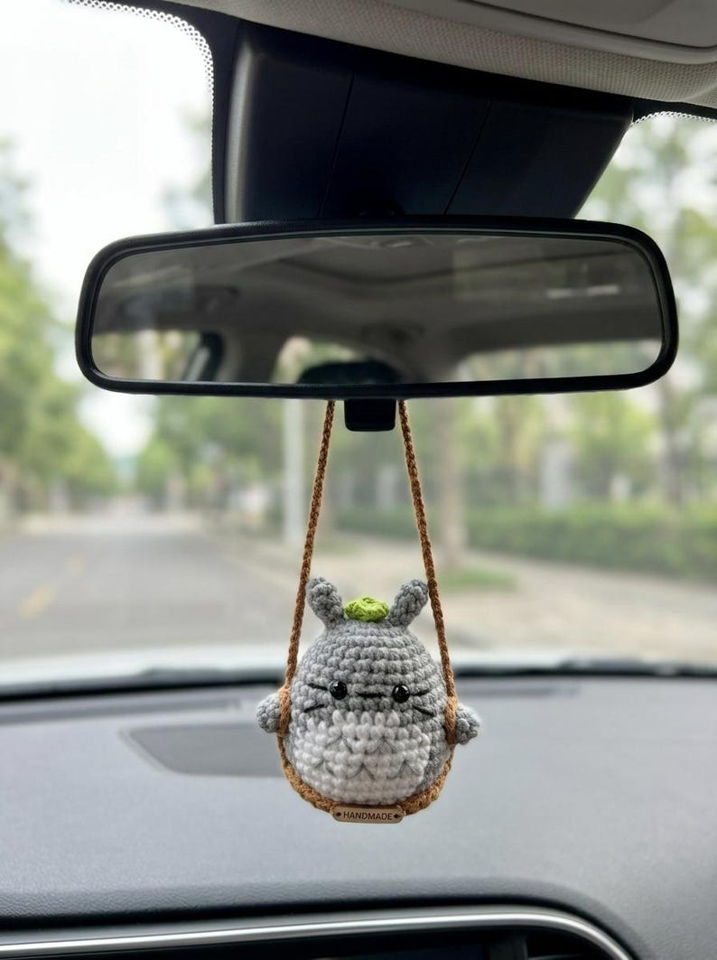 Pu&ograve; includere: Un ornamento all'uncinetto grigio e bianco appeso a un cordino marrone, con piccole orecchie, occhi neri e un germoglio verde sulla testa. La figura &egrave; seduta su un'altalena all'uncinetto marrone con una piccola etichetta rettangolare con scritto HANDMADE.