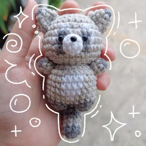 Może przedstawiać: Ręcznie robiona szydełkowa zabawka szopa w odcieniach szarości, bieli i czerni. Mała pluszowa zabawka ma okrągły korpus, pasiasty ogon i białą pierś. Szop jest trzymany w dłoni, z białymi bazgrołami wokół.