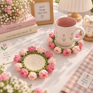 Puede incluir: Dos posavasos redondos de ganchillo con centro color crema, borde verde y un anillo de pequeñas flores tridimensionales en tonos rosa y rosa claro alrededor. Uno de los posavasos sostiene una taza de cerámica blanca con interior rosa.