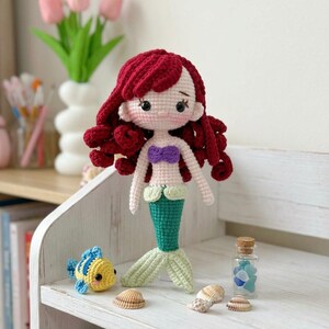 Peut inclure: Une poup&eacute;e sir&egrave;ne au crochet faite &agrave; la main avec des cheveux roux, un haut violet et une queue verte. Un poisson au crochet jaune et bleu est assis &agrave; proximit&eacute;. La poup&eacute;e est expos&eacute;e sur une &eacute;tag&egrave;re blanche avec des coquillages et une petite bouteille en verre.