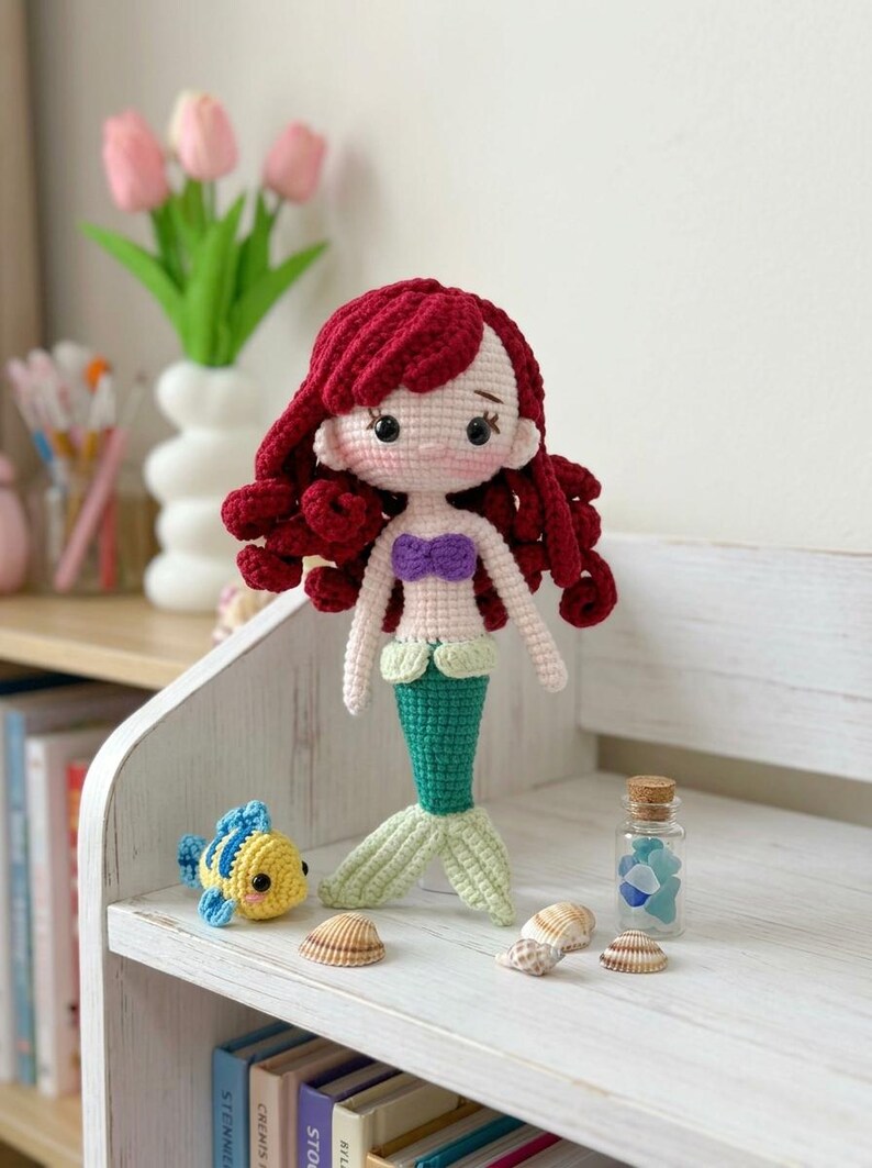 Peut inclure: Une poup&eacute;e sir&egrave;ne au crochet faite &agrave; la main avec des cheveux roux, un haut violet et une queue verte. Un poisson au crochet jaune et bleu est assis &agrave; proximit&eacute;. La poup&eacute;e est expos&eacute;e sur une &eacute;tag&egrave;re blanche avec des coquillages et une petite bouteille en verre.