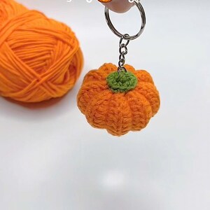 Puede incluir: Un llavero de calabaza naranja hecho a mano con ganchillo y un tallo verde. La palabra "Pumpkin" est&aacute; escrita encima del llavero. Una madeja de hilo naranja est&aacute; en el fondo. El llavero est&aacute; unido a un llavero plateado.