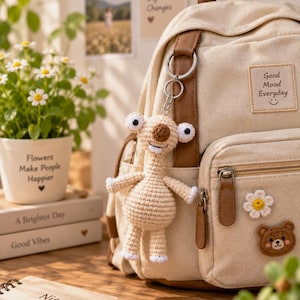 Op de afbeelding: Een beige canvas rugzak met bruine accenten in lederlook, een rechthoekige patch met de tekst Good Mood Everyday en een gehaakte luiaard-sleutelhanger. Het voorvak is voorzien van een klein madeliefje en een bruine berenpatch.