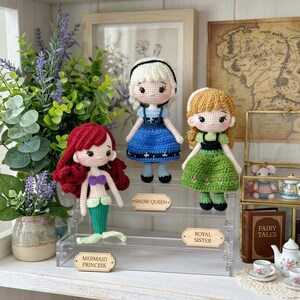Peut inclure: Trois poup&eacute;es crochet&eacute;es, une sir&egrave;ne, une reine des neiges et une s&oelig;ur royale, sont expos&eacute;es sur une &eacute;tag&egrave;re en acrylique transparent. La sir&egrave;ne a des cheveux rouges et une queue verte. La reine des neiges porte une robe bleue, et la s&oelig;ur royale porte une robe verte. Chaque poup&eacute;e a une &eacute;tiquette.