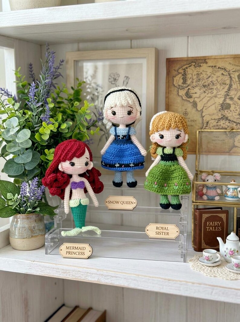 Peut inclure: Trois poup&eacute;es crochet&eacute;es, une sir&egrave;ne, une reine des neiges et une s&oelig;ur royale, sont expos&eacute;es sur une &eacute;tag&egrave;re en acrylique transparent. La sir&egrave;ne a des cheveux rouges et une queue verte. La reine des neiges porte une robe bleue, et la s&oelig;ur royale porte une robe verte. Chaque poup&eacute;e a une &eacute;tiquette.