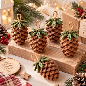 May include: Five crocheted pinecone ornaments in brown yarn with green tops, each featuring a small loop for hanging. One wooden tag displays the text "Small Creations Brighter Holidays" with a heart, and another tag reads "Handmade with Love" with a heart.