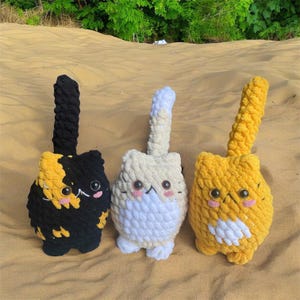 Pode incluir: Três brinquedos de gato de crochê. Um é preto e amarelo, outro é branco e amarelo, e o último é amarelo e branco.