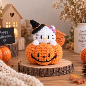 May include: A white crocheted cat figure with a purple bow, wearing a black witch hat with an orange band, sitting inside a round orange crocheted pumpkin with a black stitched jack-o'-lantern face and an orange tag reading Handmade with Love.