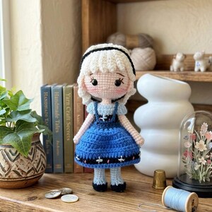 Peut inclure: Une poup&eacute;e au crochet faite &agrave; la main, v&ecirc;tue d'une robe bleue, de cheveux blancs et d'un bandeau noir. La poup&eacute;e a une expression douce et se tient sur une surface en bois. Un bel exemple d'artisanat.