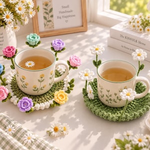 Op de afbeelding: Twee keramische mokken met bloemmotieven, elk rustend op een ronde groene gehaakte onderzetter. De onderzetters zijn versierd met driedimensionale gehaakte bloemen in diverse kleuren, waaronder roze, blauw, geel, paars en witte madeliefjes.