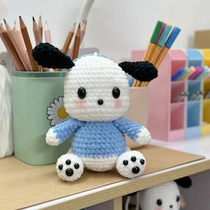 Peut inclure: Une poupée chien au crochet faite à la main, avec une tête et un corps blancs, un pull bleu et des oreilles noires. La poupée a des yeux et des pattes noirs. Elle est posée sur une étagère en bois avec des crayons et des stylos en arrière-plan.