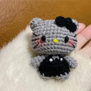 Peut inclure: Une figurine de chat au crochet faite &agrave; la main, avec une t&ecirc;te grise, un n&oelig;ud noir et une robe noire. Le chat a des yeux noirs, des joues roses et un nez jaune. La figurine est pos&eacute;e sur une surface blanche et duveteuse.