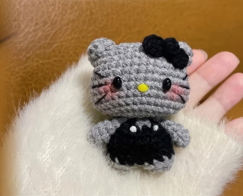 Peut inclure: Une figurine de chat au crochet faite &agrave; la main, avec une t&ecirc;te grise, un n&oelig;ud noir et une robe noire. Le chat a des yeux noirs, des joues roses et un nez jaune. La figurine est pos&eacute;e sur une surface blanche et duveteuse.