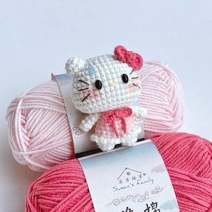 Peut inclure: Une poup&eacute;e chat au crochet avec un n&oelig;ud rose et une robe, plac&eacute;e sur deux pelotes de fil rose. Le fil porte une &eacute;tiquette avec du texte chinois et les mots "Susan's Family".