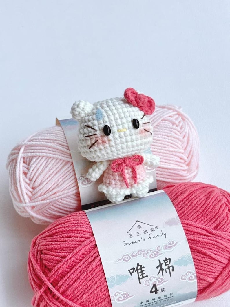Peut inclure: Une poup&eacute;e chat au crochet avec un n&oelig;ud rose et une robe, plac&eacute;e sur deux pelotes de fil rose. Le fil porte une &eacute;tiquette avec du texte chinois et les mots "Susan's Family".