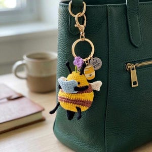 May include: A handmade crochet bee keychain with yellow and brown stripes, wearing a blue mask. The keychain includes a gold ring, a purple flower, and a charm that says "BEE KIND". The bee is attached to a dark green leather handbag.