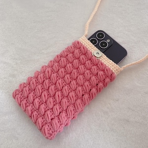Puede incluir: Una funda de teléfono rosa tejida a ganchillo con un patrón texturizado en forma de burbujas. La funda tiene una correa rosa claro y un borde beige. Un teléfono inteligente es visible dentro de la funda, que se cierra con un broche plateado.