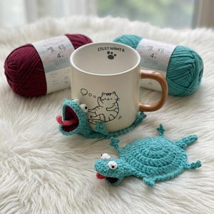 May include: A cream-colored mug with a brown handle and a cat illustration, resting on a turquoise crochet frog coaster. Two skeins of yarn, one burgundy and one teal, are in the background. The mug reads "I JUST WANT A".