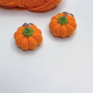 Puede incluir: Dos llaveros de calabaza naranja de ganchillo con tallos verdes sobre una superficie blanca. Una madeja de hilo naranja y la palabra "Pumpkin" est&aacute;n en el fondo. Los llaveros est&aacute;n hechos a mano.