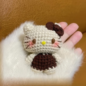 Peut inclure: Une figurine Hello Kitty au crochet de couleur cr&egrave;me, marron et rose. La figurine a des yeux en boutons marron, un nez jaune et un n&oelig;ud marron. La figurine porte une robe marron et a des marques roses sur les joues.