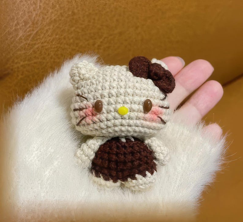 Peut inclure: Une figurine Hello Kitty au crochet de couleur cr&egrave;me, marron et rose. La figurine a des yeux en boutons marron, un nez jaune et un n&oelig;ud marron. La figurine porte une robe marron et a des marques roses sur les joues.